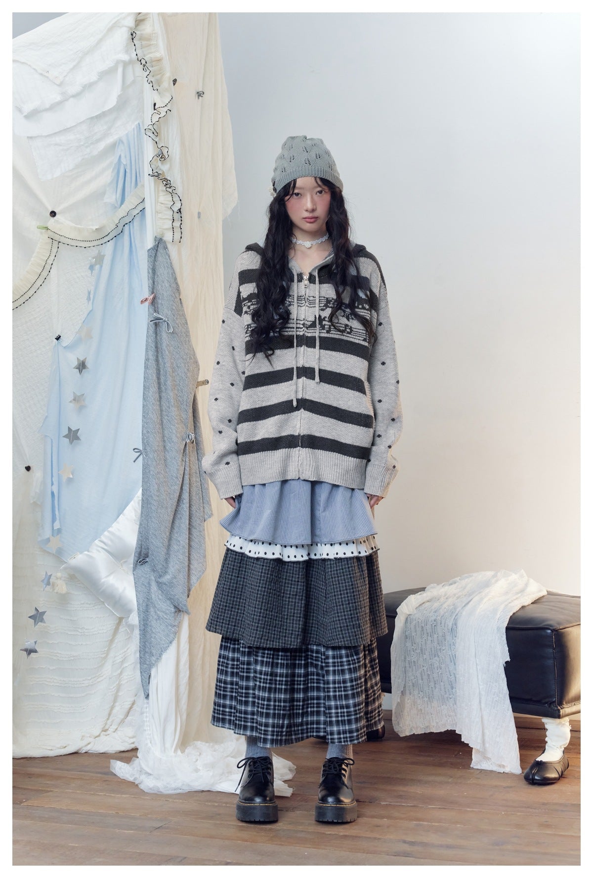 Model wearing gray striped zip hooded knit cardigan with dotted sleeves over a layered plaid skirt, front view