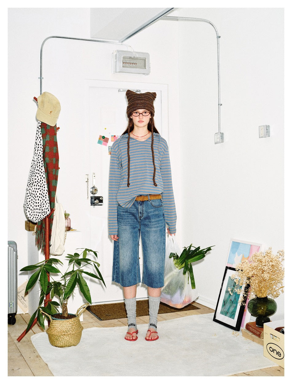Woman wearing a blue and taupe striped long-sleeve top with jeans and a knit hat, standing in a white room
