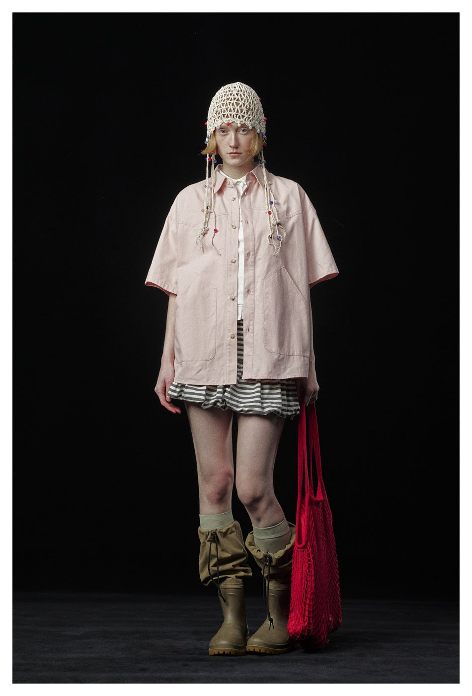 Model wearing the pink short-sleeve shirt open over a white top, with striped shorts, a crochet beaded cap and boots