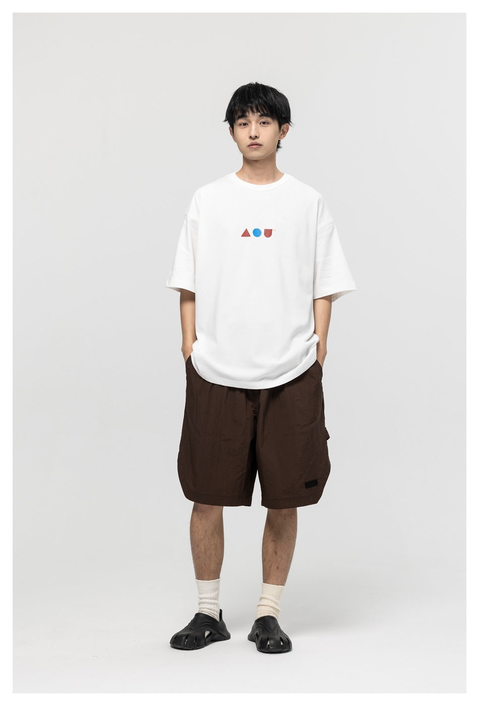 Full-length model wearing a white oversized graphic T-shirt, dark brown shorts, white socks, and black sandals.