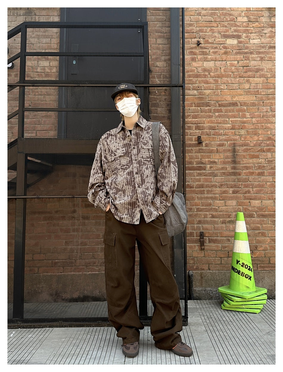 Person wearing brown striped tie-dye-style shirt with brown cargo pants and a cap, standing by a brick wall