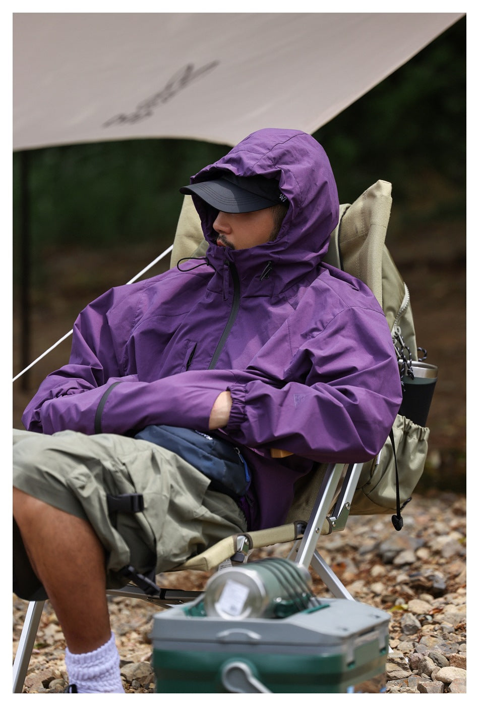 Person seated outdoors wearing a purple hooded jacket with a black cap.
