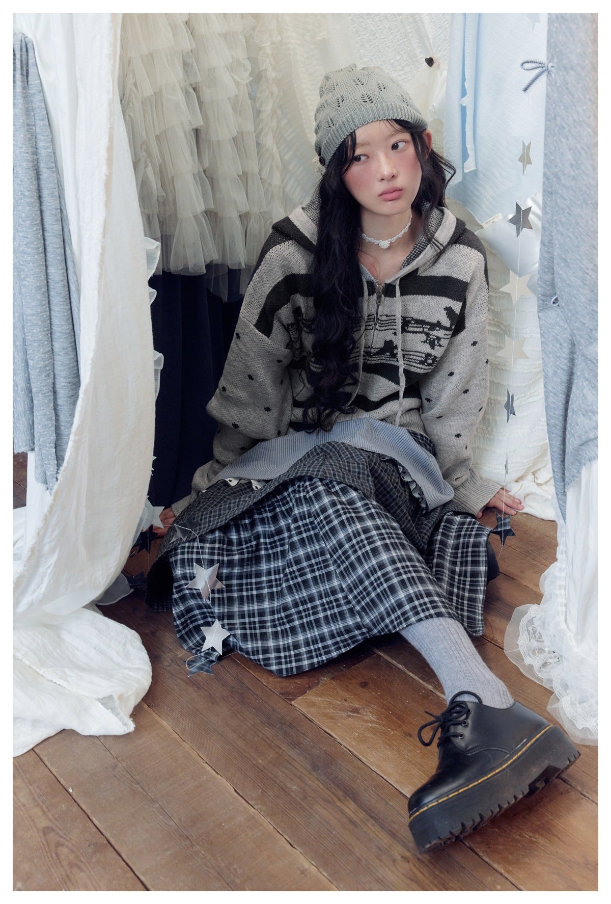 Seated model wearing a gray and black patterned hooded sweater with a long dark plaid skirt.