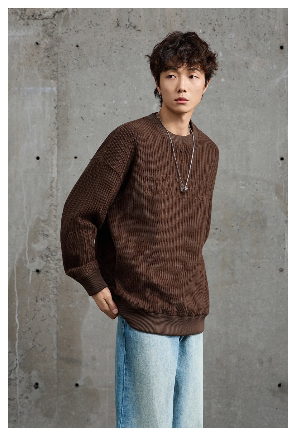 Angled view of a model in a dark brown textured crewneck sweater with subtle tonal chest lettering and light-wash jeans.