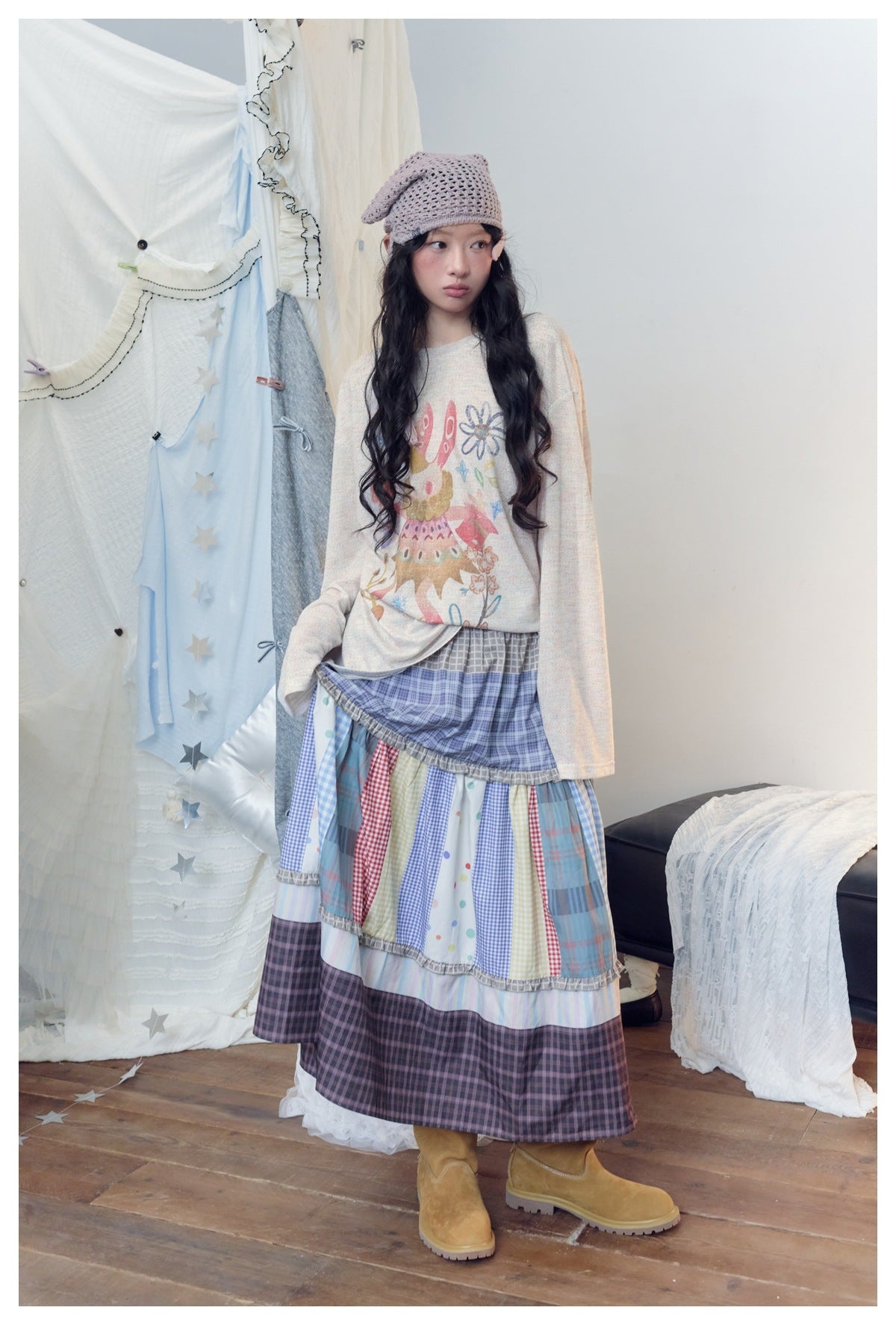Model wearing a cream illustrated long-sleeve top, a blue and multicolor tiered patchwork skirt, a mauve knit cap and tan