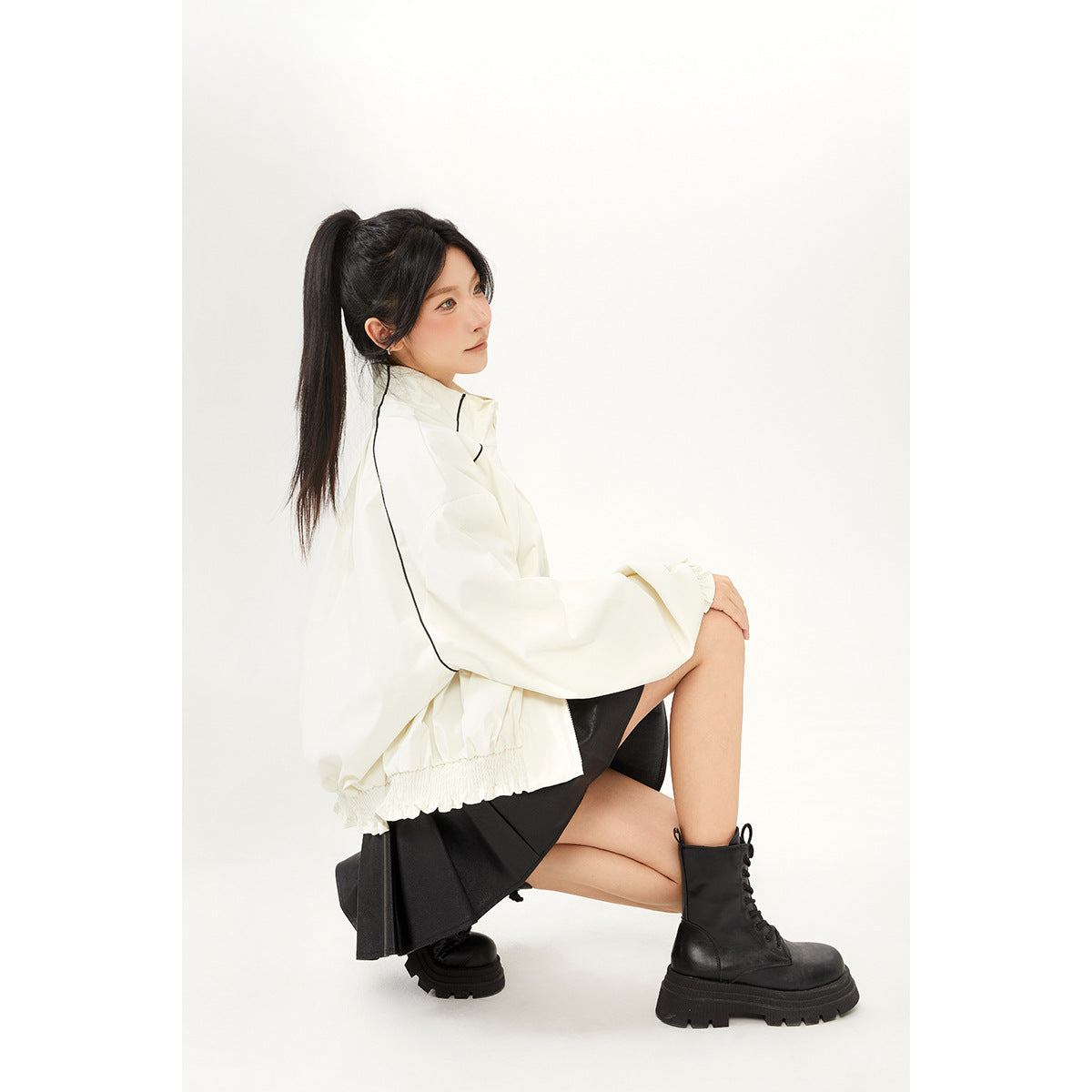 Seated side view of a model in an ivory jacket with black piping, a black skirt, and chunky lace-up boots.