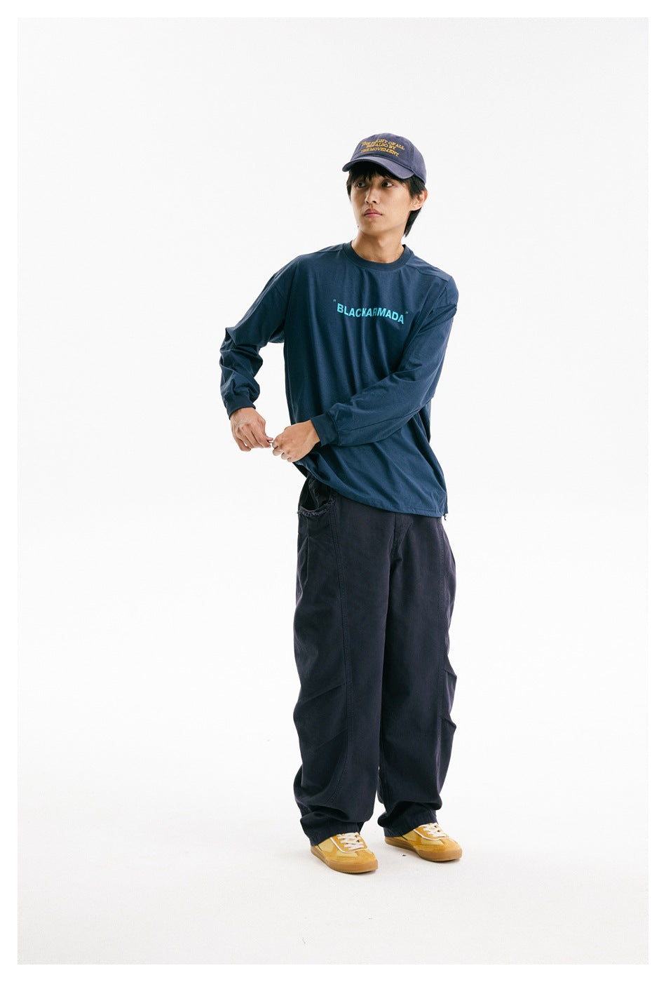 Model wearing a navy long-sleeve shirt with cyan chest lettering and loose dark pants, adjusting the hem.