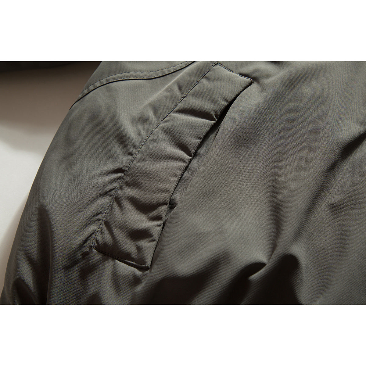 Close-up of a dark olive-gray jacket showing a stitched flap pocket and gathered fabric.