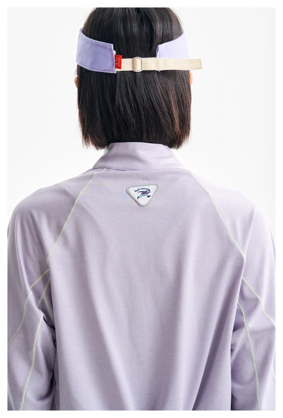 Close-up of the lilac top's back seams and small badge, with the adjustable strap of a lavender visor visible.
