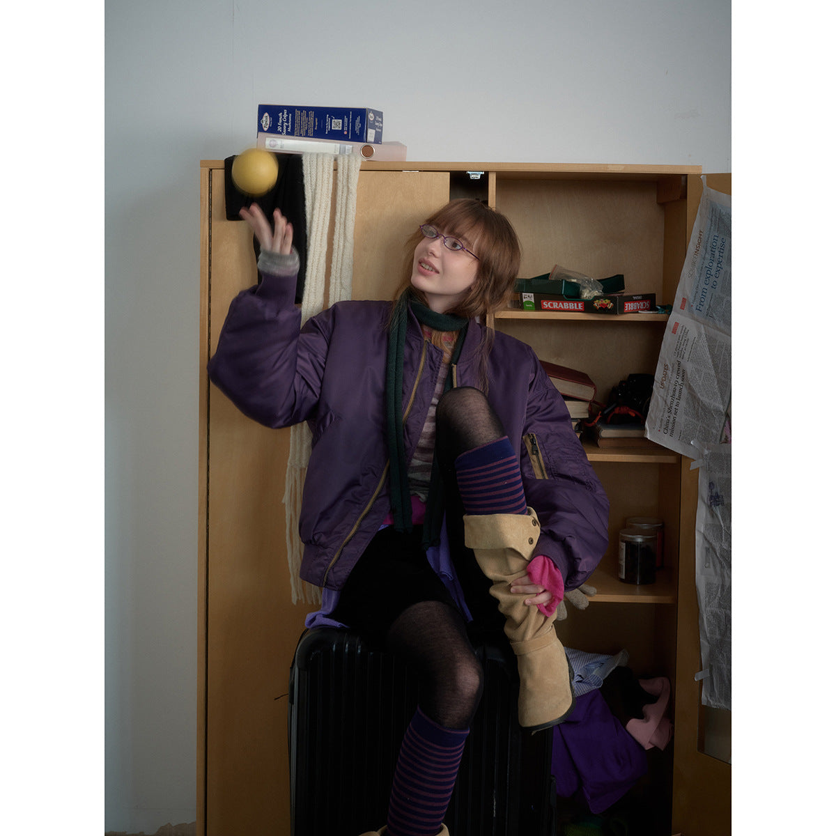 Model in a purple bomber jacket with zip pockets, seated in a cluttered room.