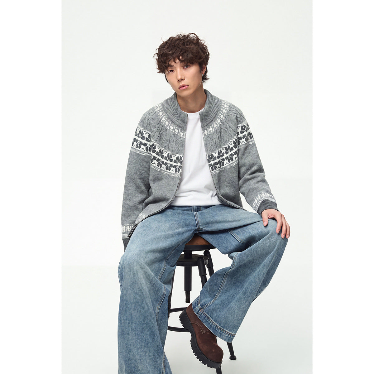 Model seated wearing a gray zip-up knit cardigan with white and dark patterned bands, a white shirt, and blue jeans.