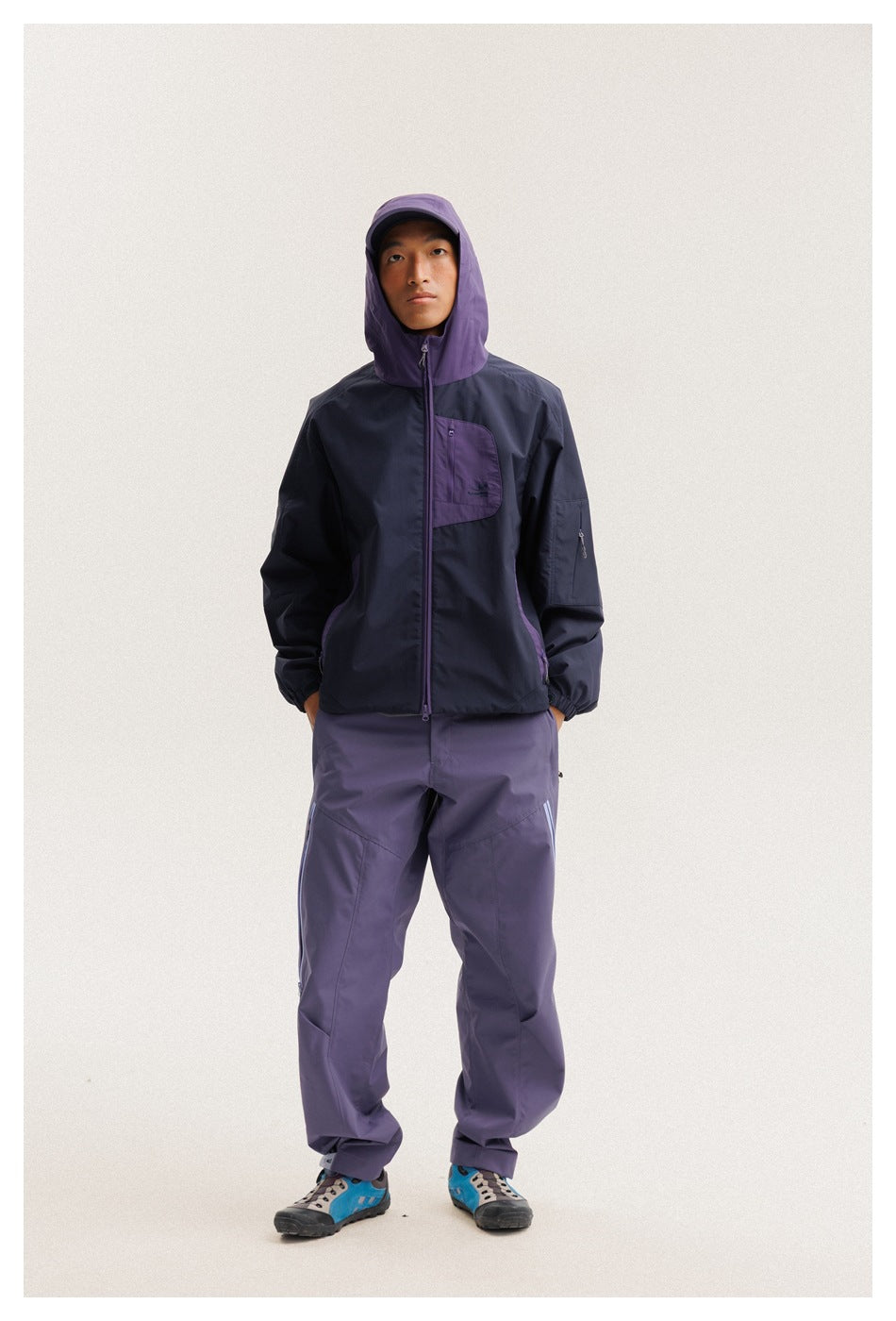 Full-length front view of a model wearing a navy jacket with its purple hood up and matching purple pants.