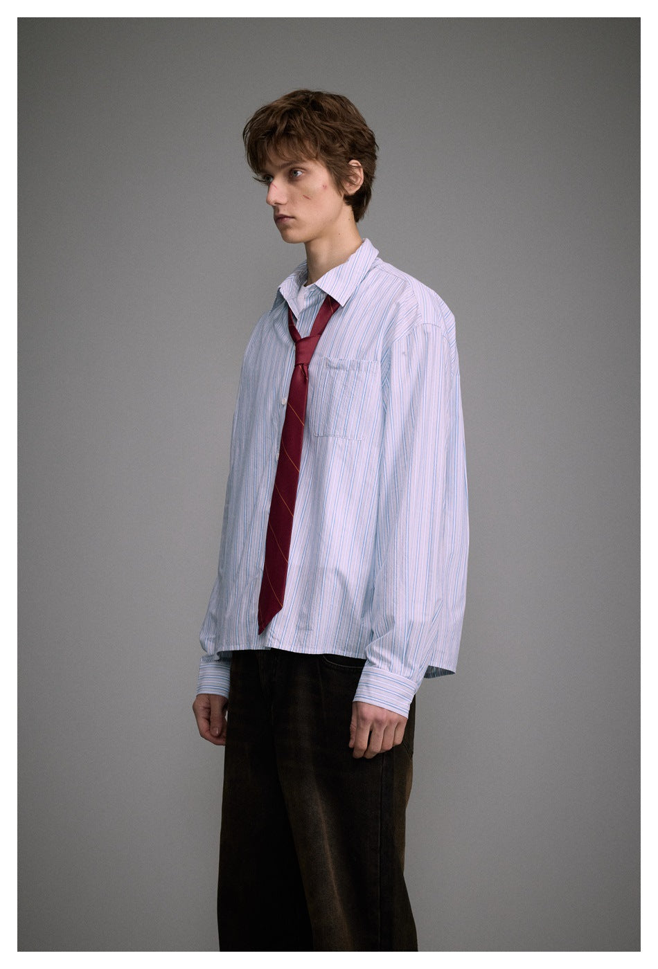 Model wearing a loose blue-and-white striped button-up shirt with a chest pocket and burgundy tie.