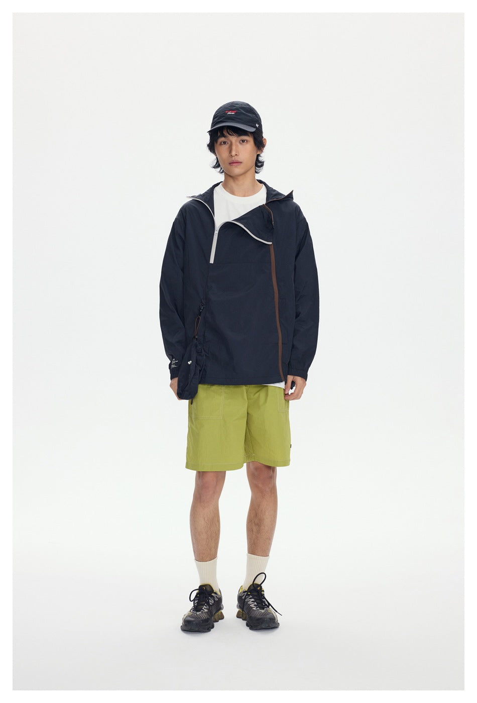 Model wearing black hooded jacket with diagonal zipper, white tee, green shorts and cap, front view