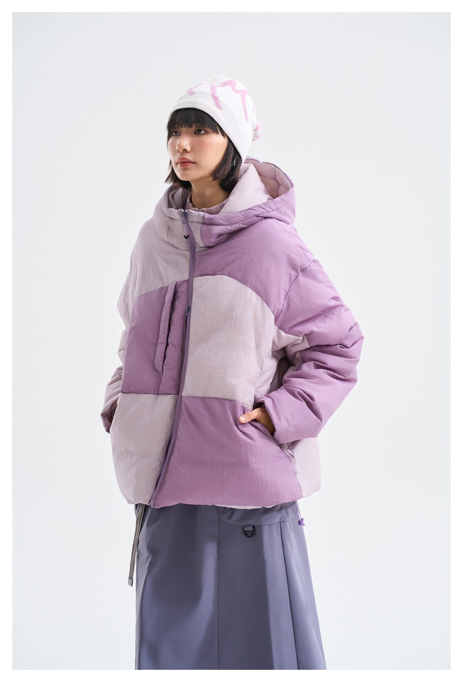 Model wearing the purple color-block hooded puffer jacket with hand in pocket, side view with a gray-purple skirt
