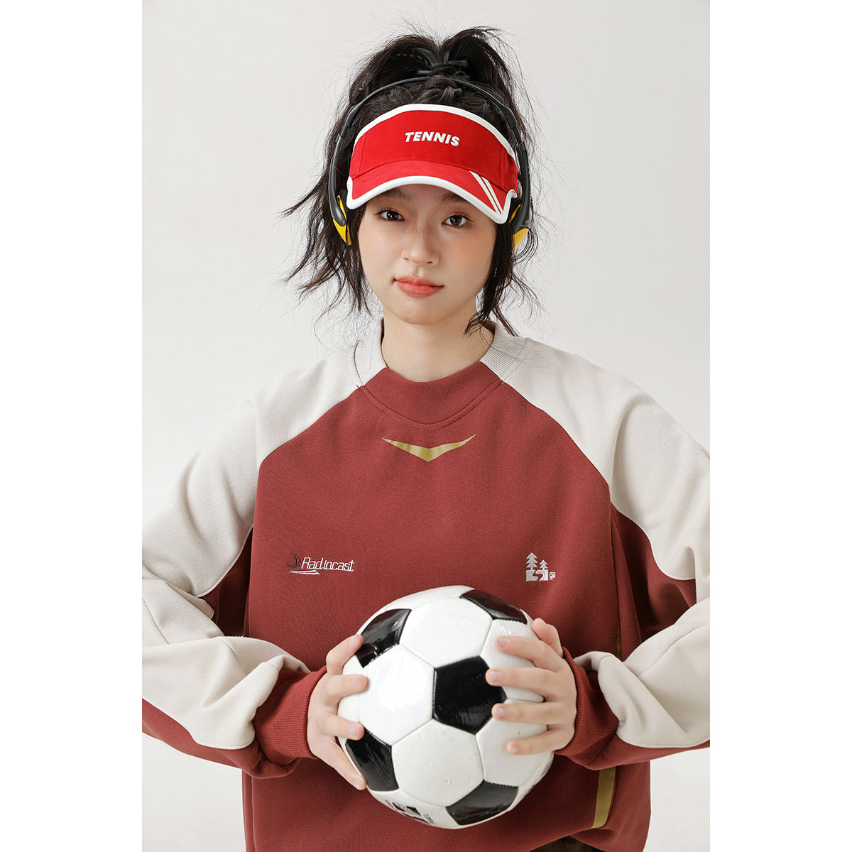 Woman wearing a rust-red and cream color-block crewneck sweatshirt while holding a soccer ball.