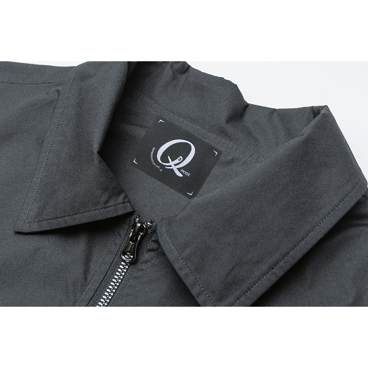 Close-up of a dark jacket's pointed collar, inner neck label, stitching, and metal front zipper.
