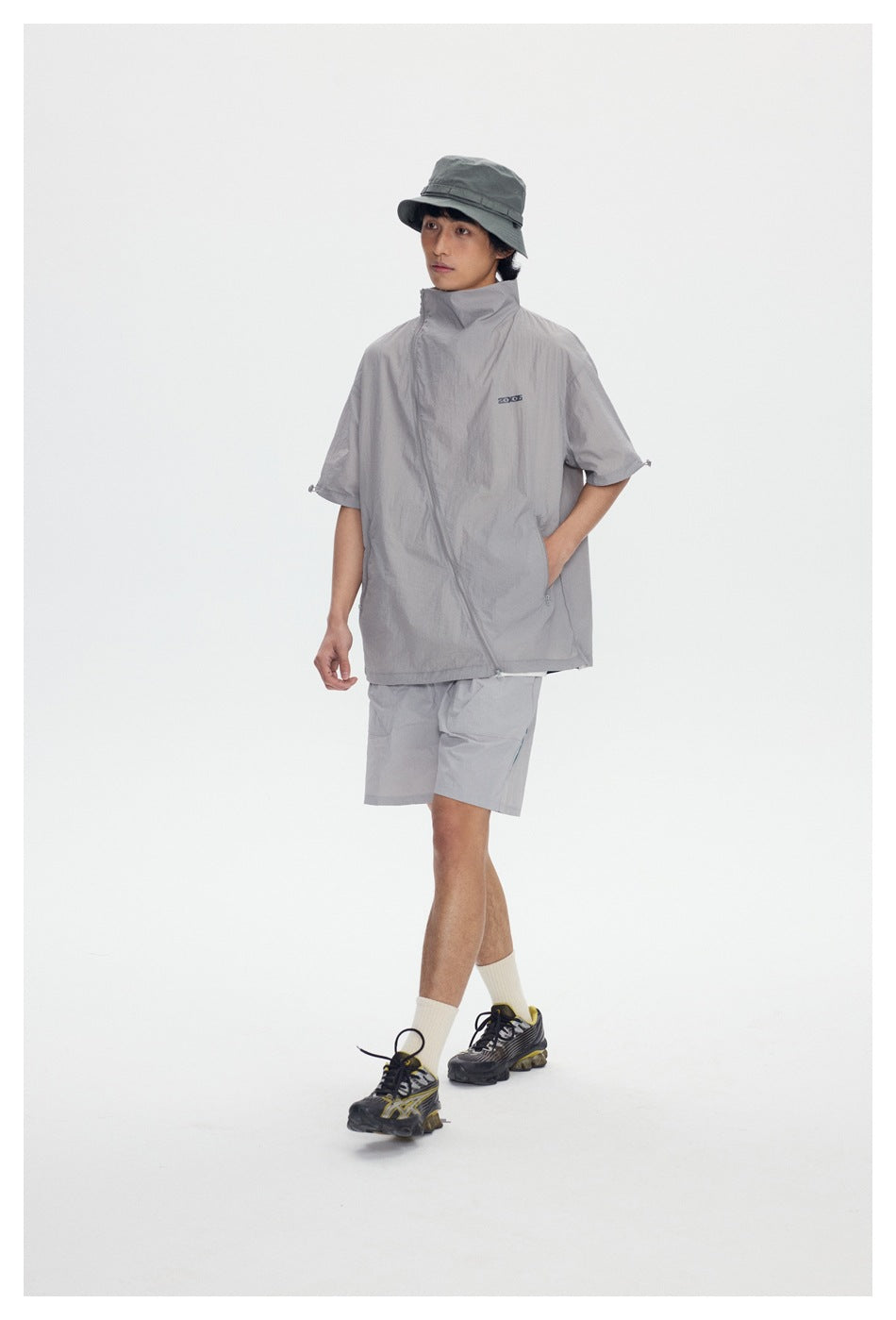 Model walking in gray short-sleeve stand-collar jacket with diagonal zipper, hand in pocket, with shorts and bucket hat