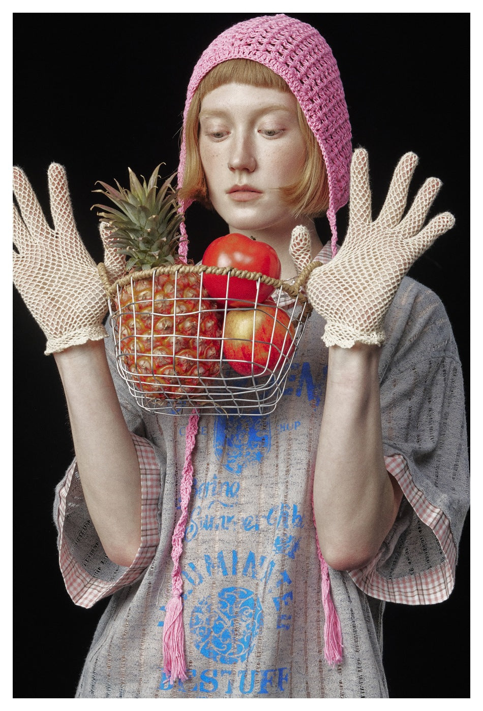 Close-up of a model wearing a gray striped blue-graphic top, pink crocheted bonnet and cream mesh gloves beside a fruit