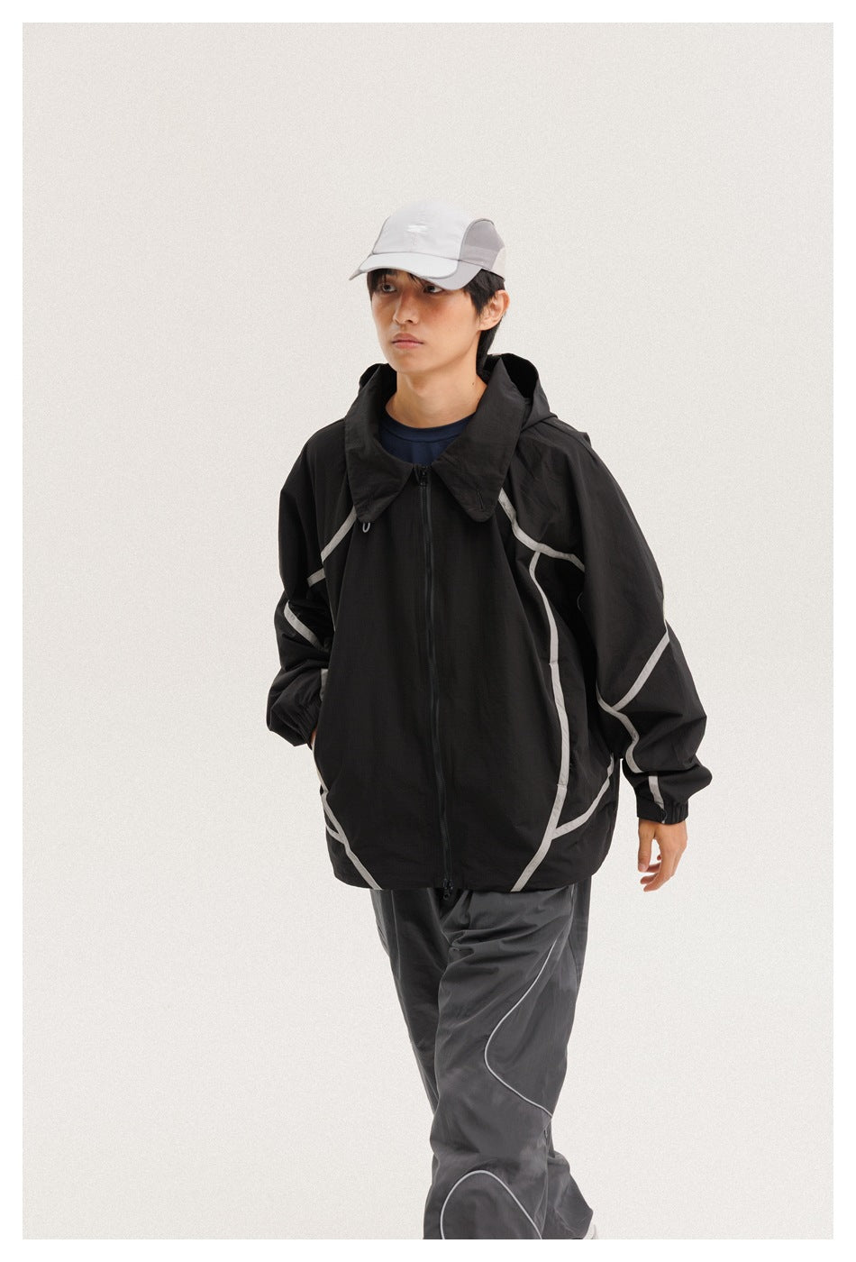 Model wearing a black zip-front hooded jacket with light curved piping, charcoal pants, and a white cap.