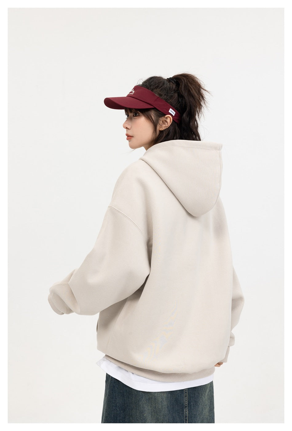 Back view of a model wearing an oversized cream hoodie with a hood and a maroon visor.