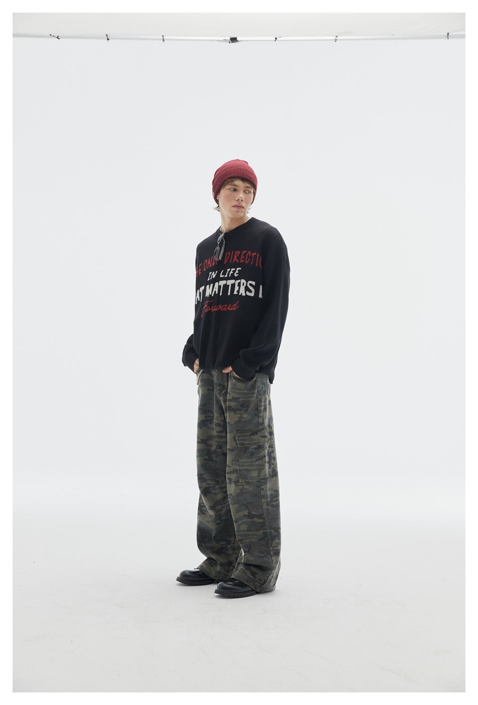 Three-quarter view of a model wearing a black lettered crewneck sweater, burgundy beanie, and camouflage cargo pants.