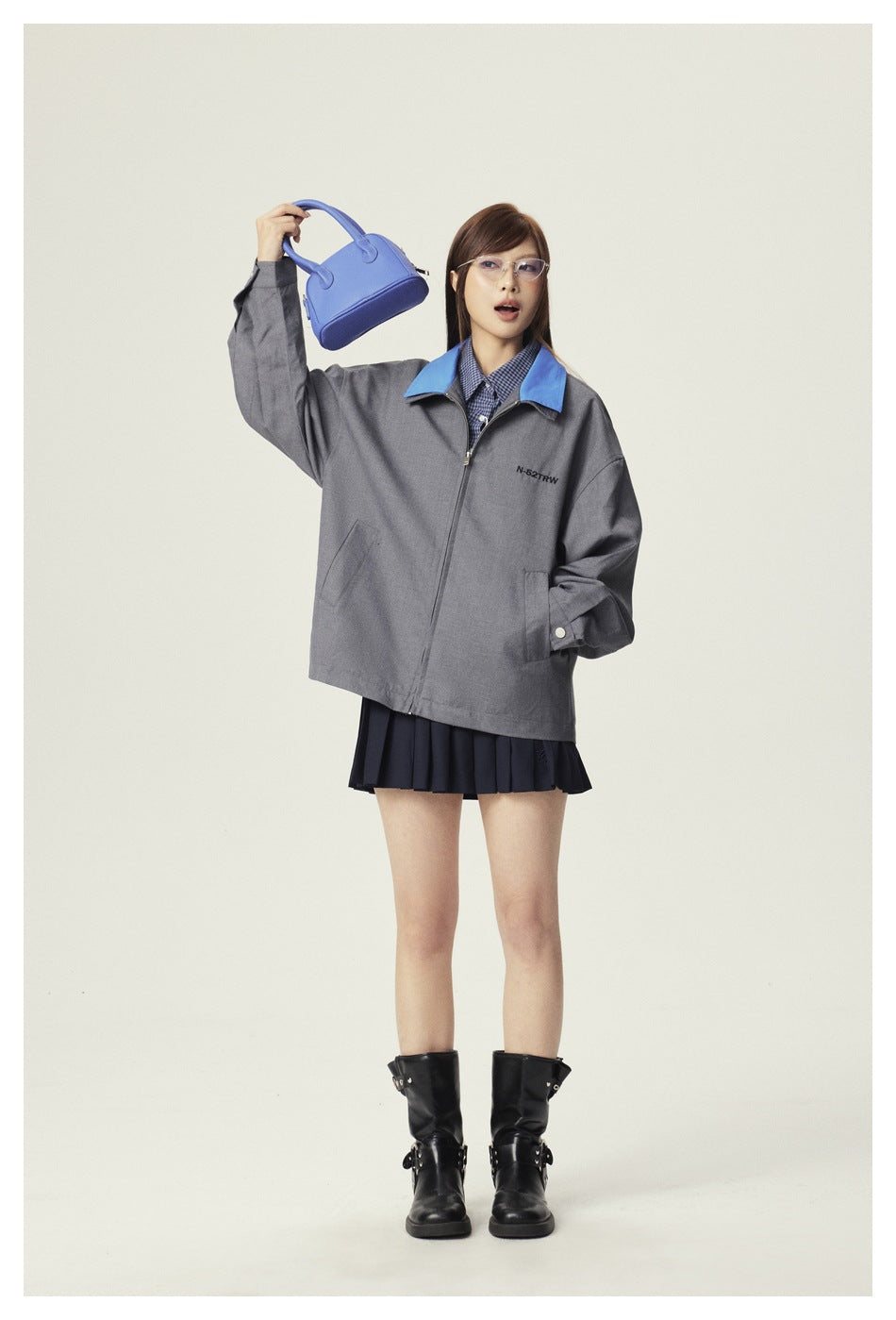 Model wearing gray zip jacket with blue collar over a checked shirt and pleated navy skirt, holding a blue handbag