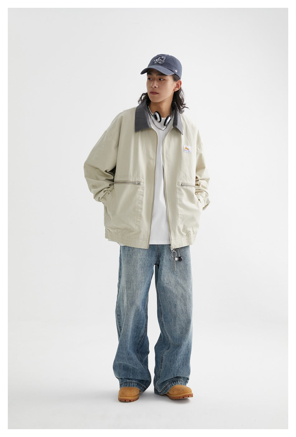 Full-length view of a model wearing a pale beige jacket with a gray collar and zip pockets, loose blue jeans, and a cap.