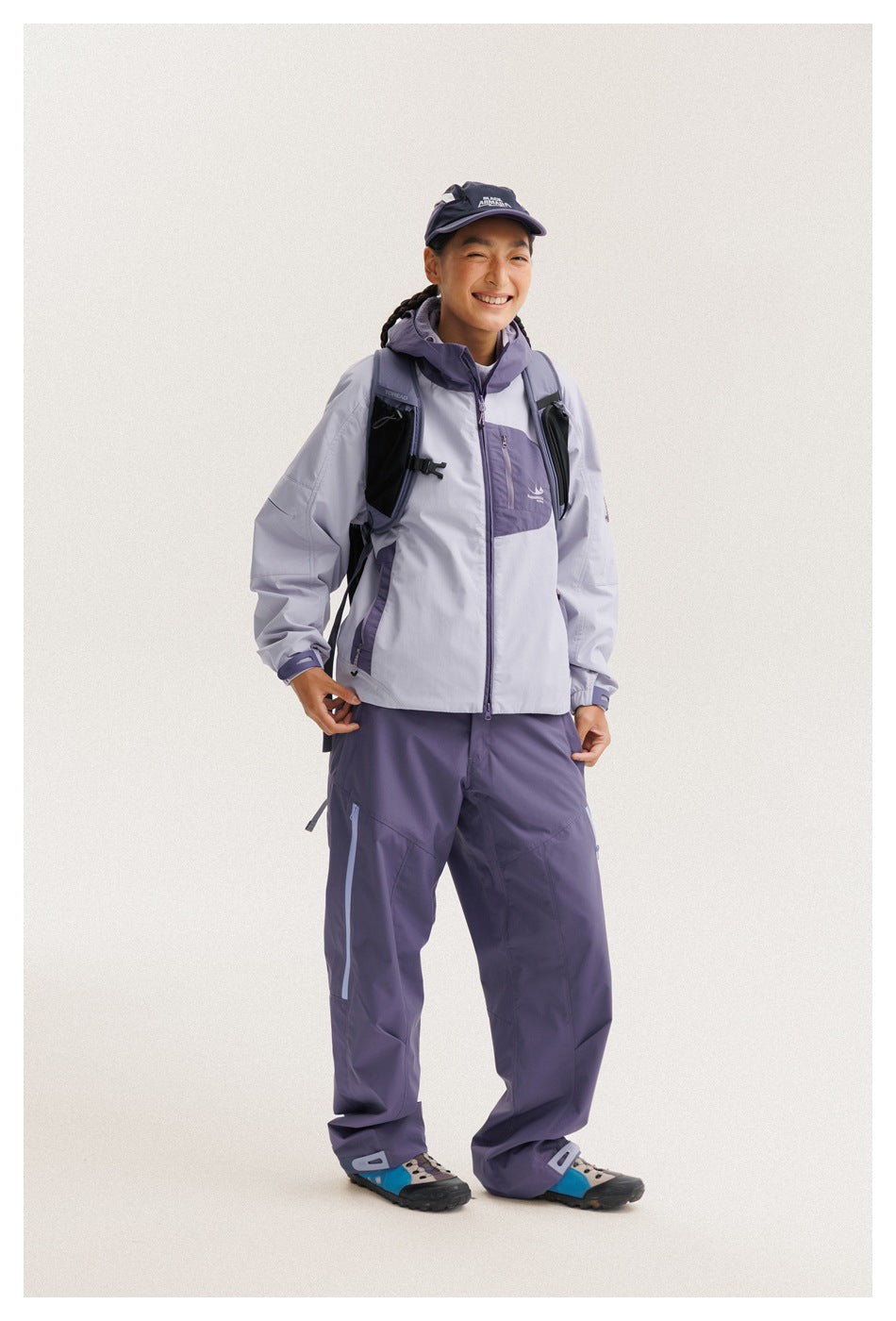 Model wearing a light gray and purple hooded jacket with matching purple trousers and a backpack.