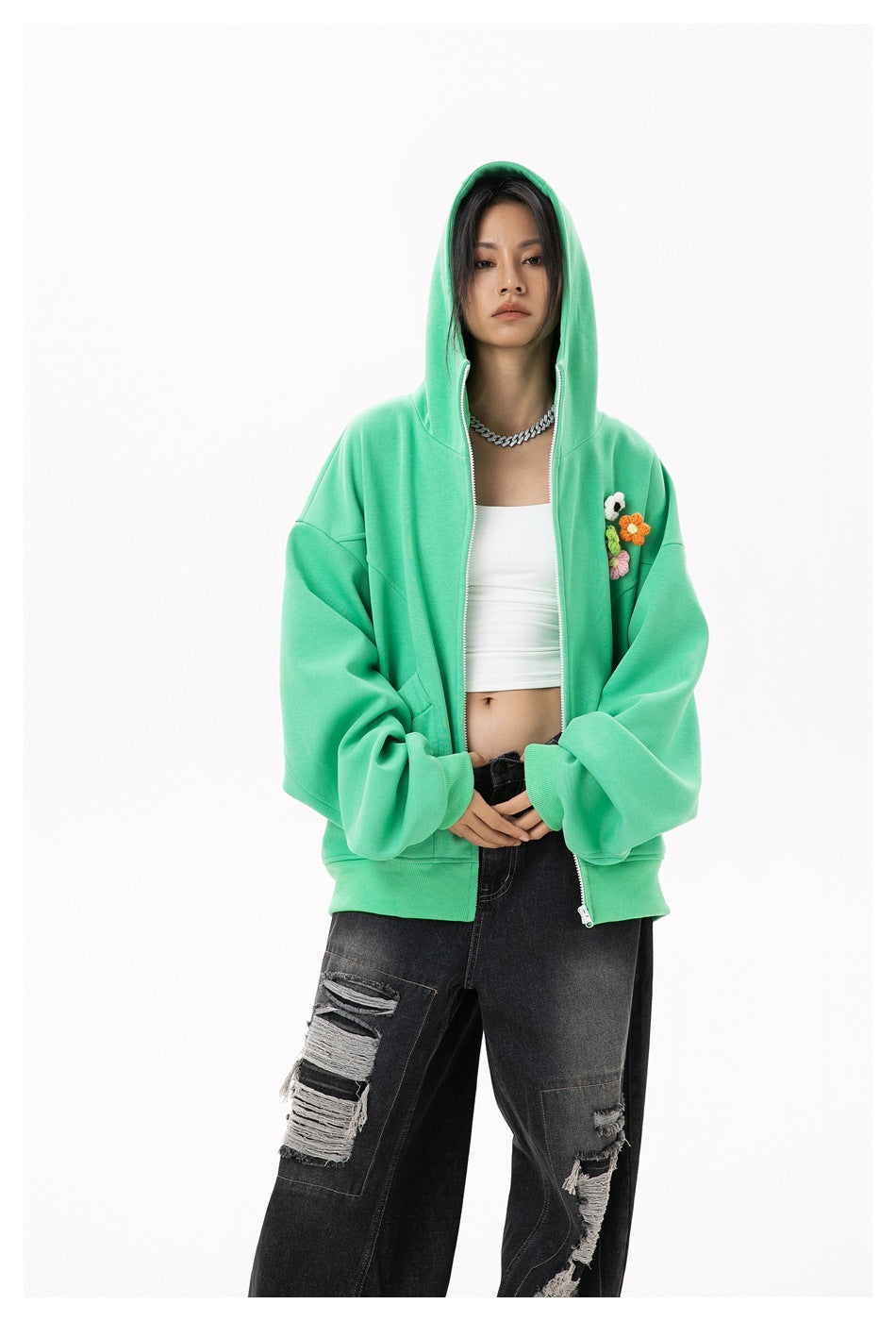 Model wearing a mint-green zip hoodie with a hood and colorful flower appliqués, styled with a white top and black jeans.