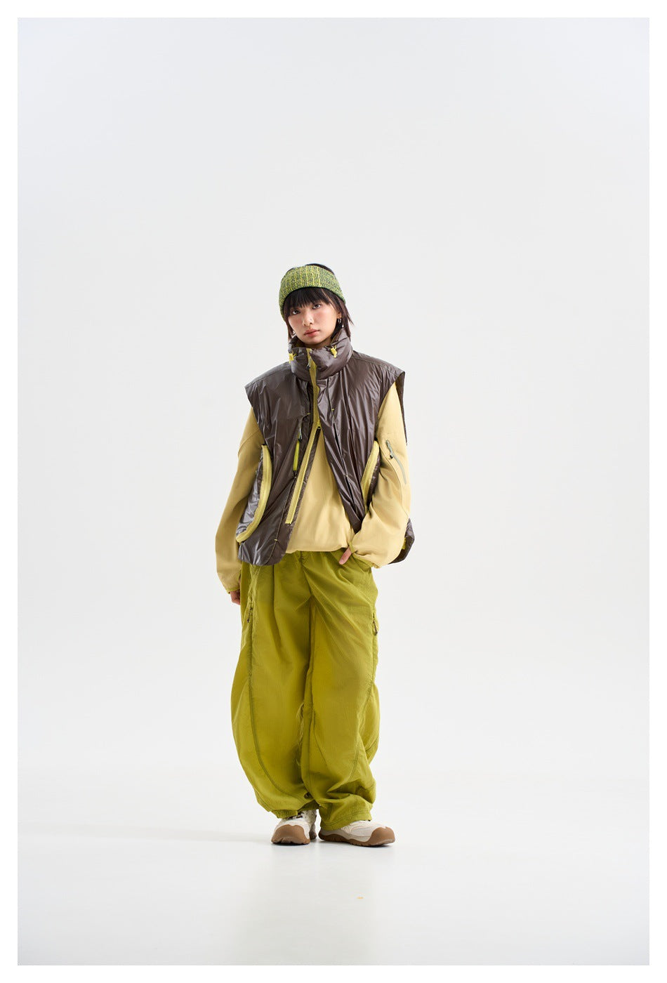 Model in a high-collar charcoal vest with yellow details, a pale yellow top and loose lime-green trousers.