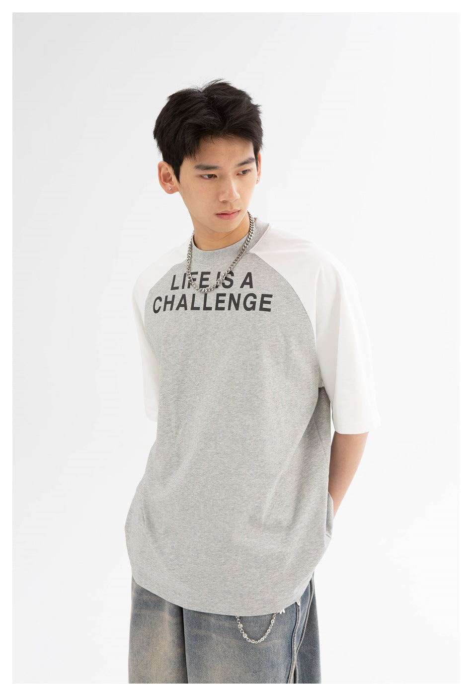 Model wearing a gray raglan T-shirt with white sleeves and bold black “LIFE IS A CHALLENGE” chest print.