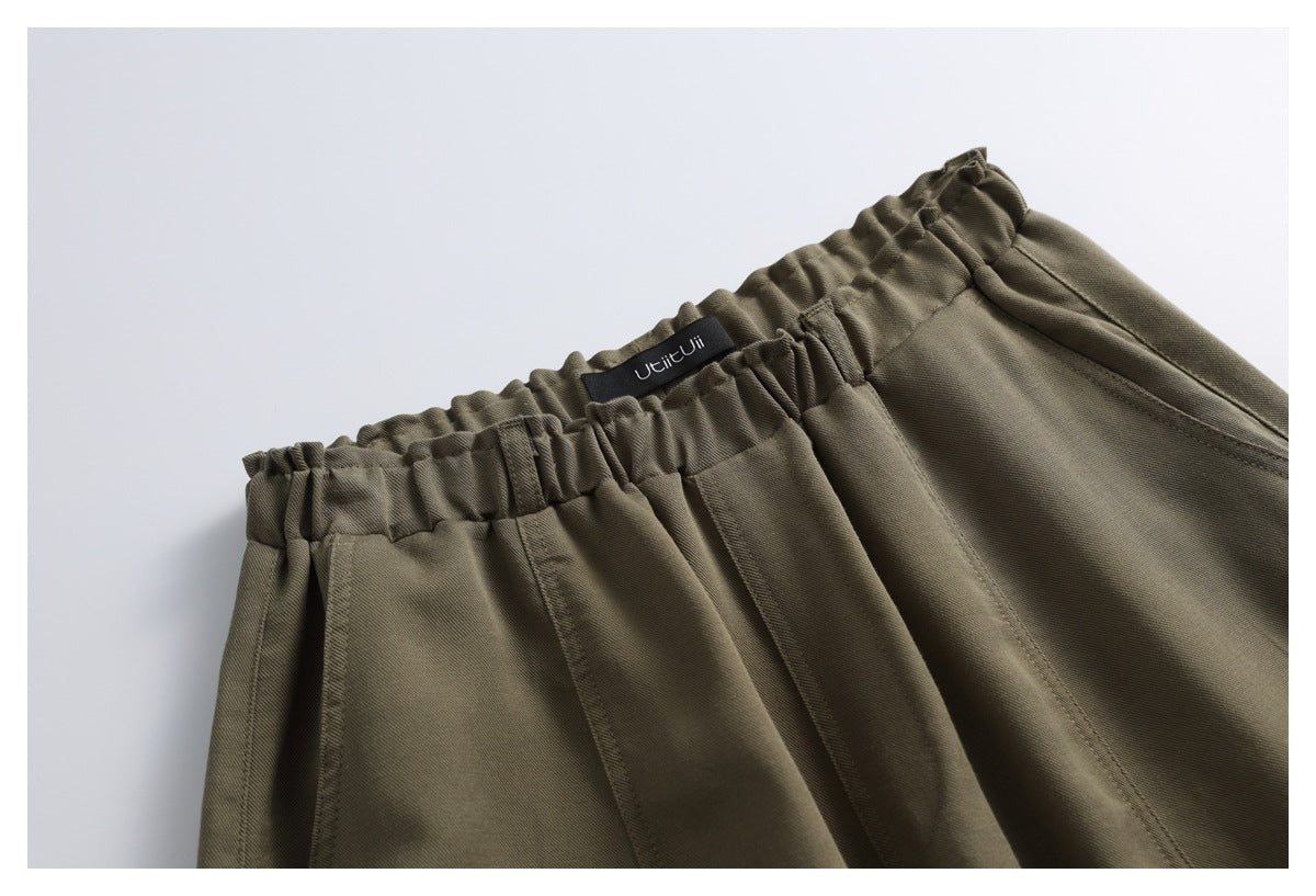 Close-up of olive-brown trousers showing a gathered elastic waistband, side pocket opening and small black waistband label.