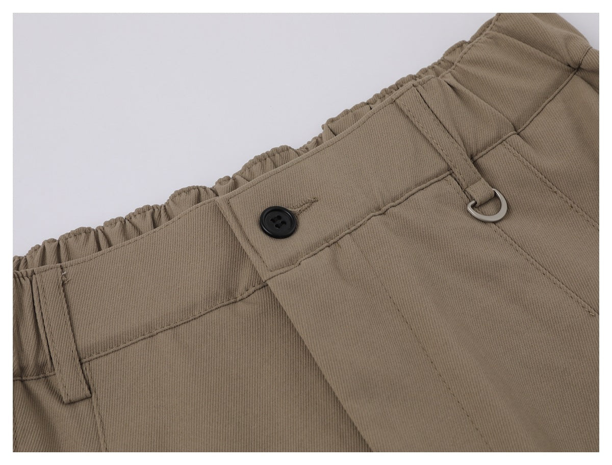 Close-up of taupe trousers with an elastic waistband, black button, belt loop, and metal D-ring.