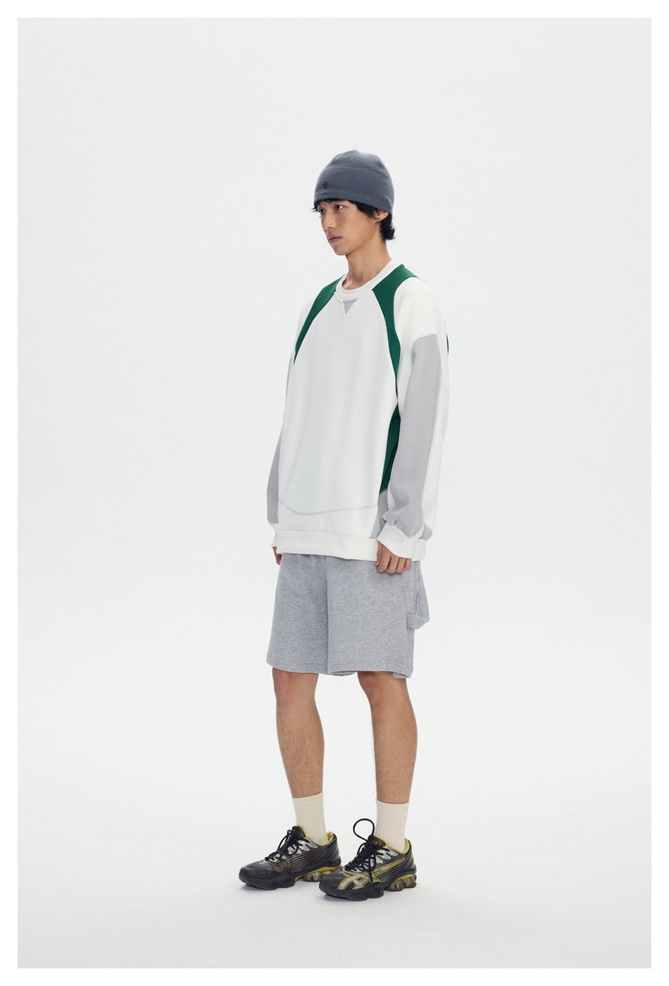 Model in gray knee-length shorts, side view, with a white and green sweatshirt, beanie and sneakers