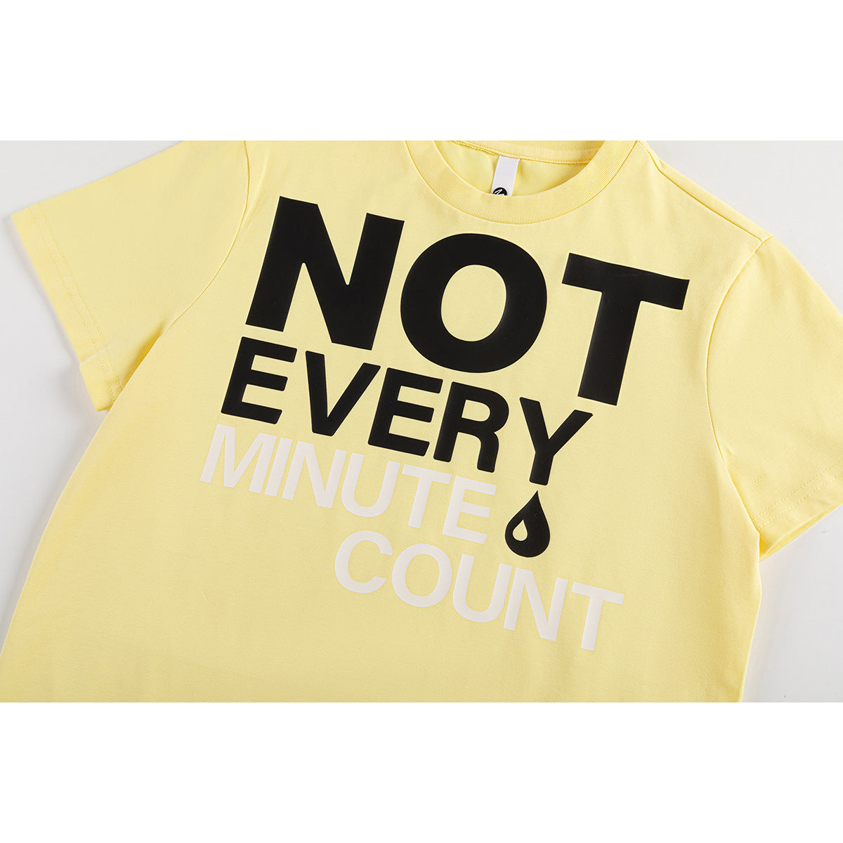 Close-up of a pale yellow T-shirt front with bold black and white “NOT EVERY MINUTE COUNT” lettering and a black droplet.