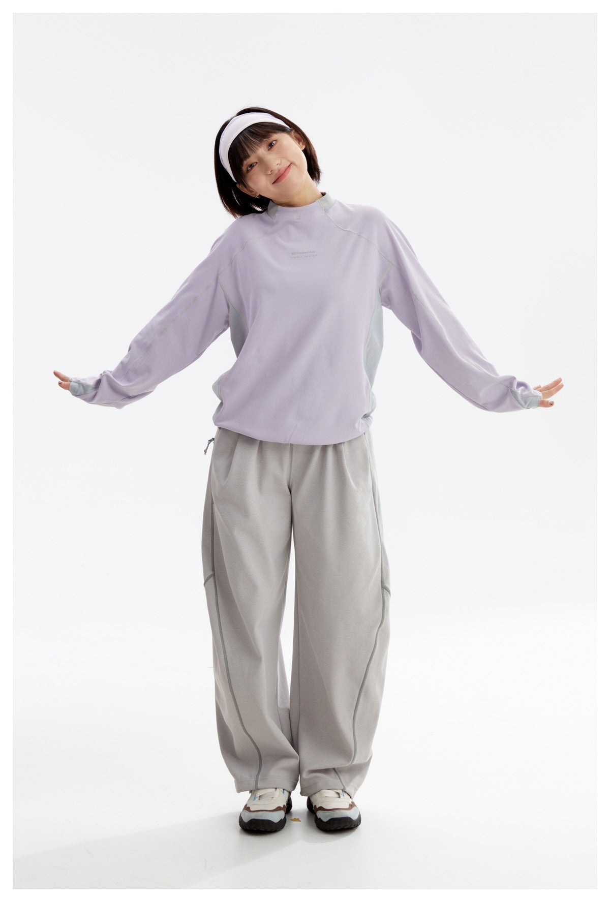 Woman wearing a lavender high-neck top and loose gray pants, standing with arms outstretched.