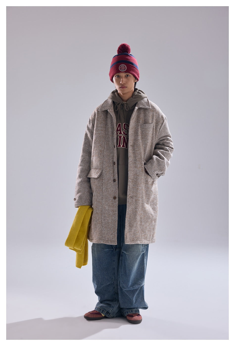 Person in khaki herringbone mid-length coat over a gray hoodie, wide jeans and a red beanie, front view