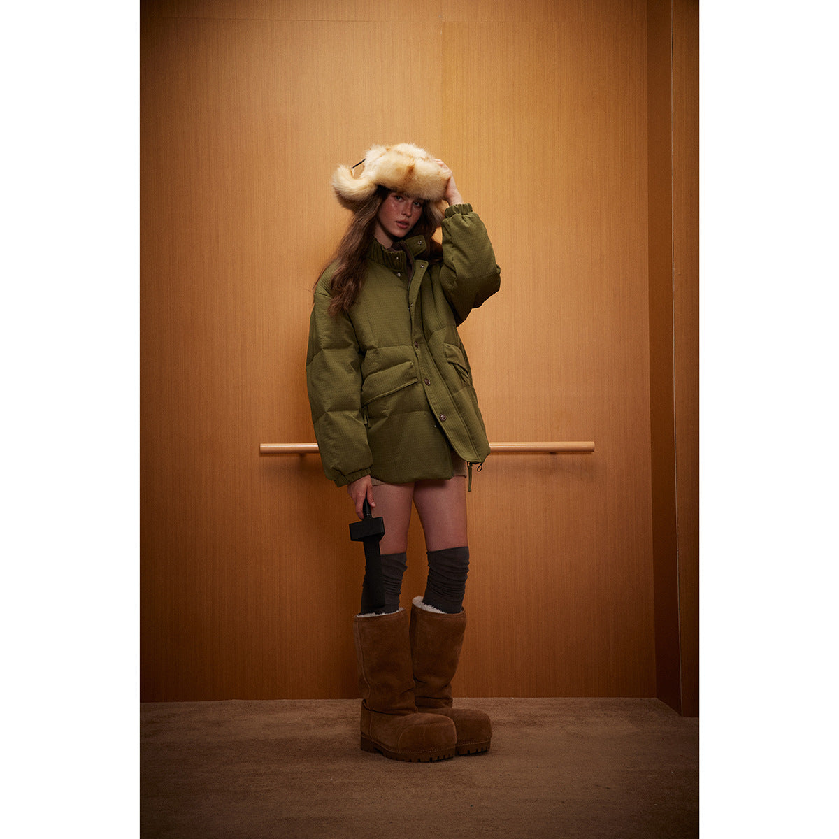 Model wearing an olive-green padded jacket with a large cream fur hat, dark leg warmers, and brown boots.