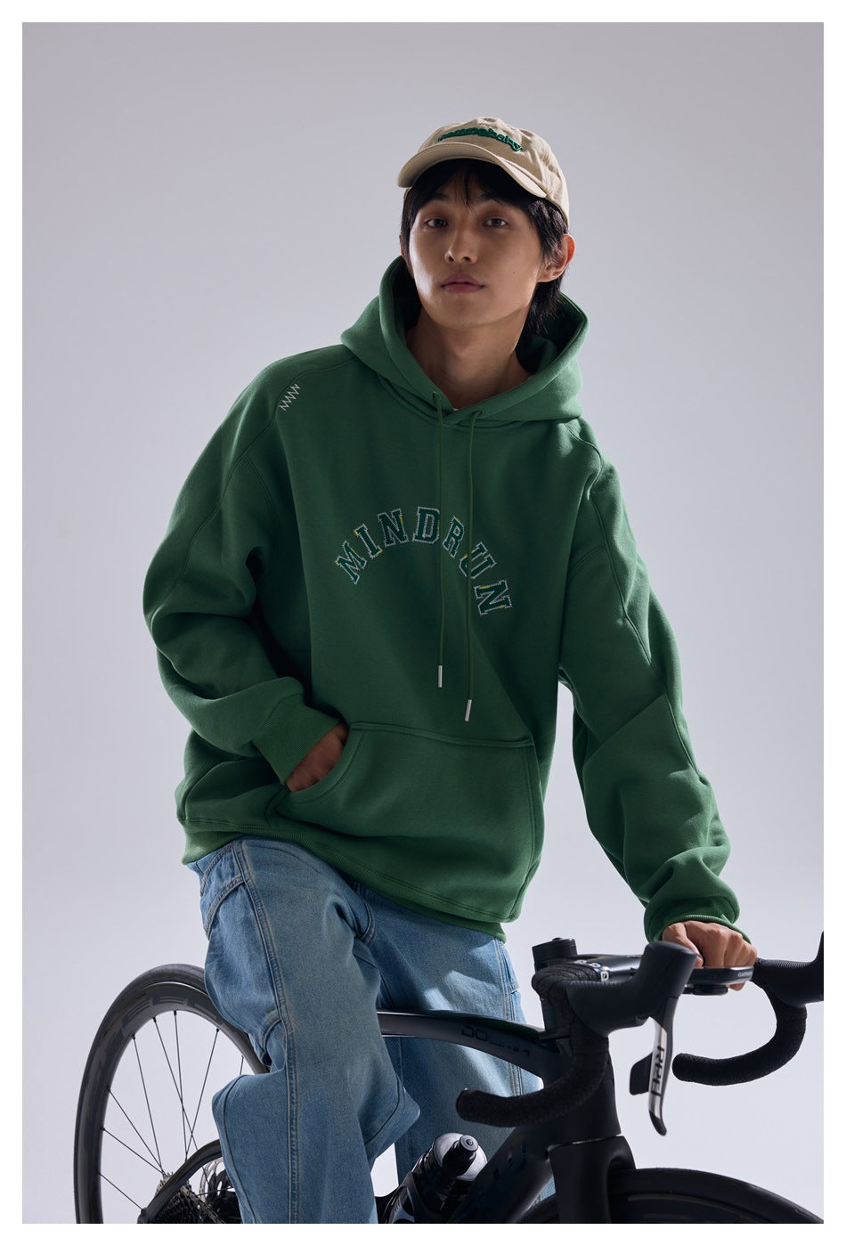 Model seated on a bicycle wearing a dark green MINDRUN hoodie with a kangaroo pocket, blue jeans and a beige cap.