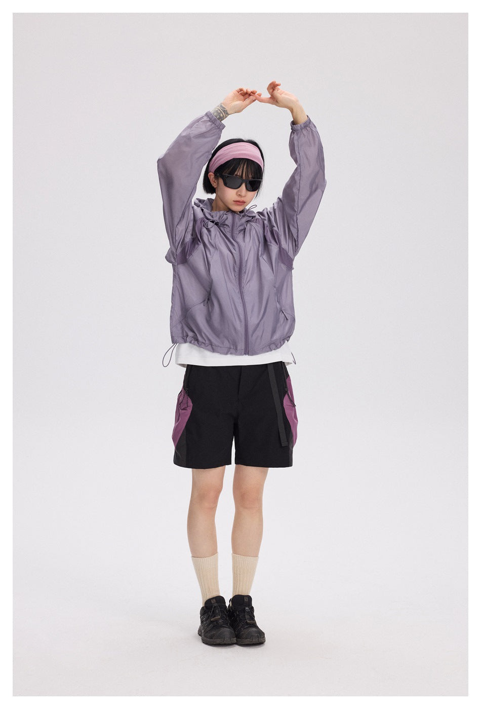 A model wears a lavender hooded jacket and black shorts with magenta side pockets, raising her arms.