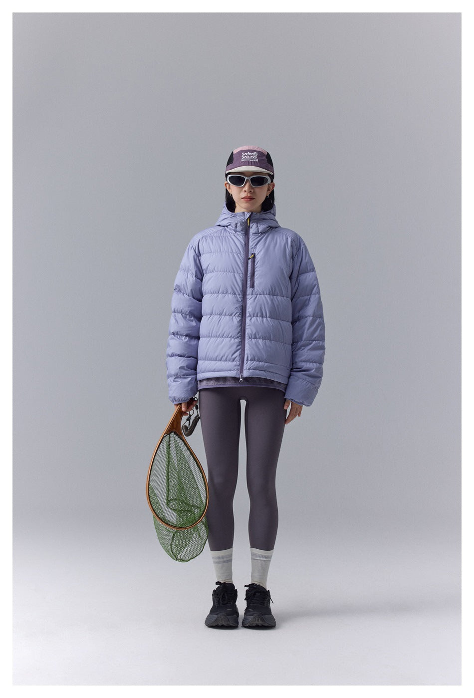 Model wearing a pale lavender quilted hooded jacket zipped up, paired with dark leggings and a cap.