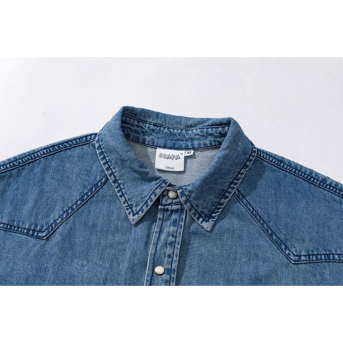 Close-up of a blue denim shirt collar, shoulder seams, snap buttons, and neck label.