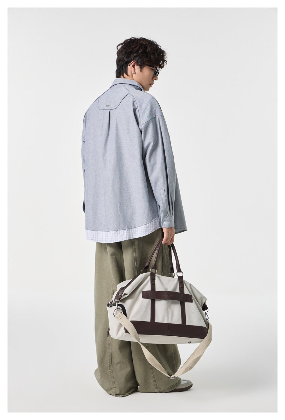 Back view of man in gray shirt with shoulder yoke and plaid hem layer, holding a canvas bag