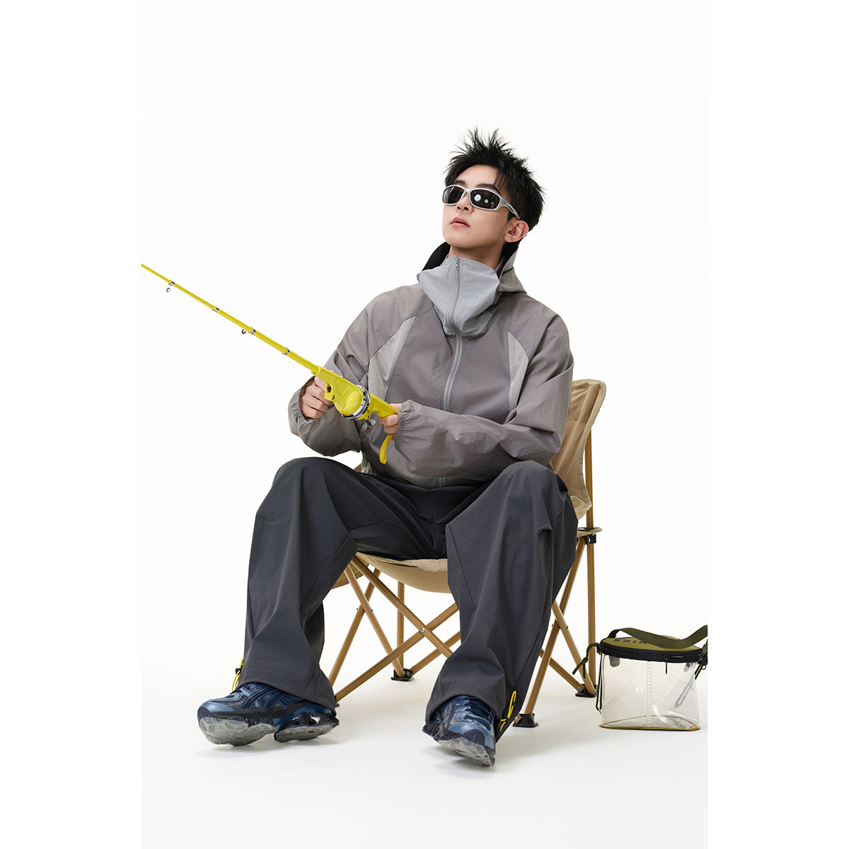 Seated model wearing a gray high-collar zip jacket and charcoal wide-leg trousers while holding a yellow fishing rod.