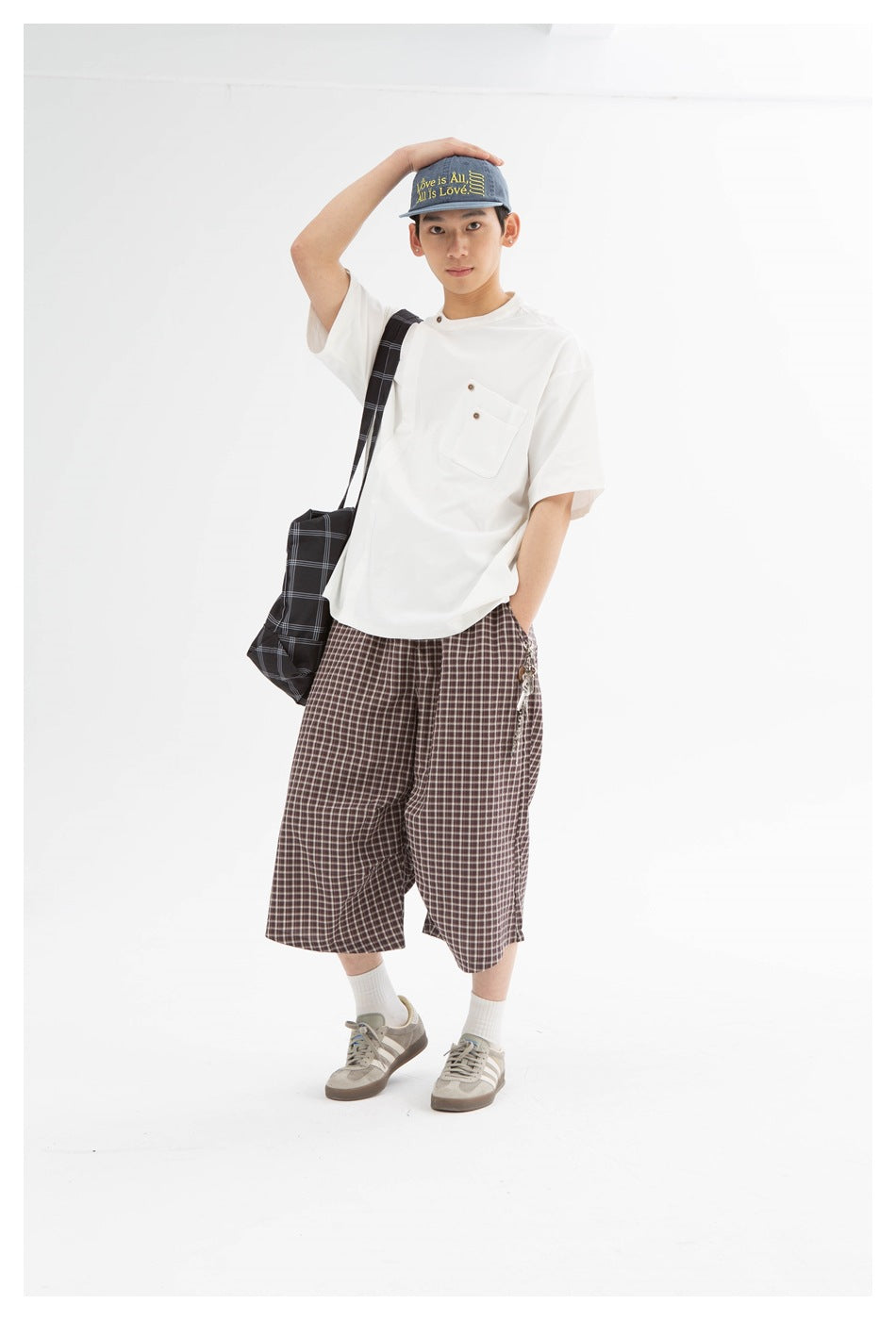 Model wearing brown plaid knee-length shorts and a white pocket T-shirt, shown from the front with a checked shoulder bag.