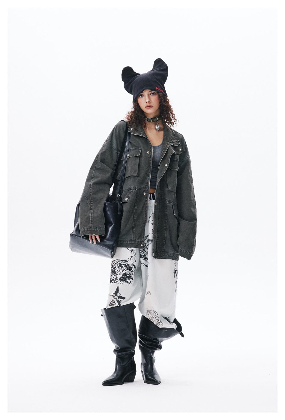 Model wearing an open dark green washed utility jacket over a gray tank top, with printed pants and black boots