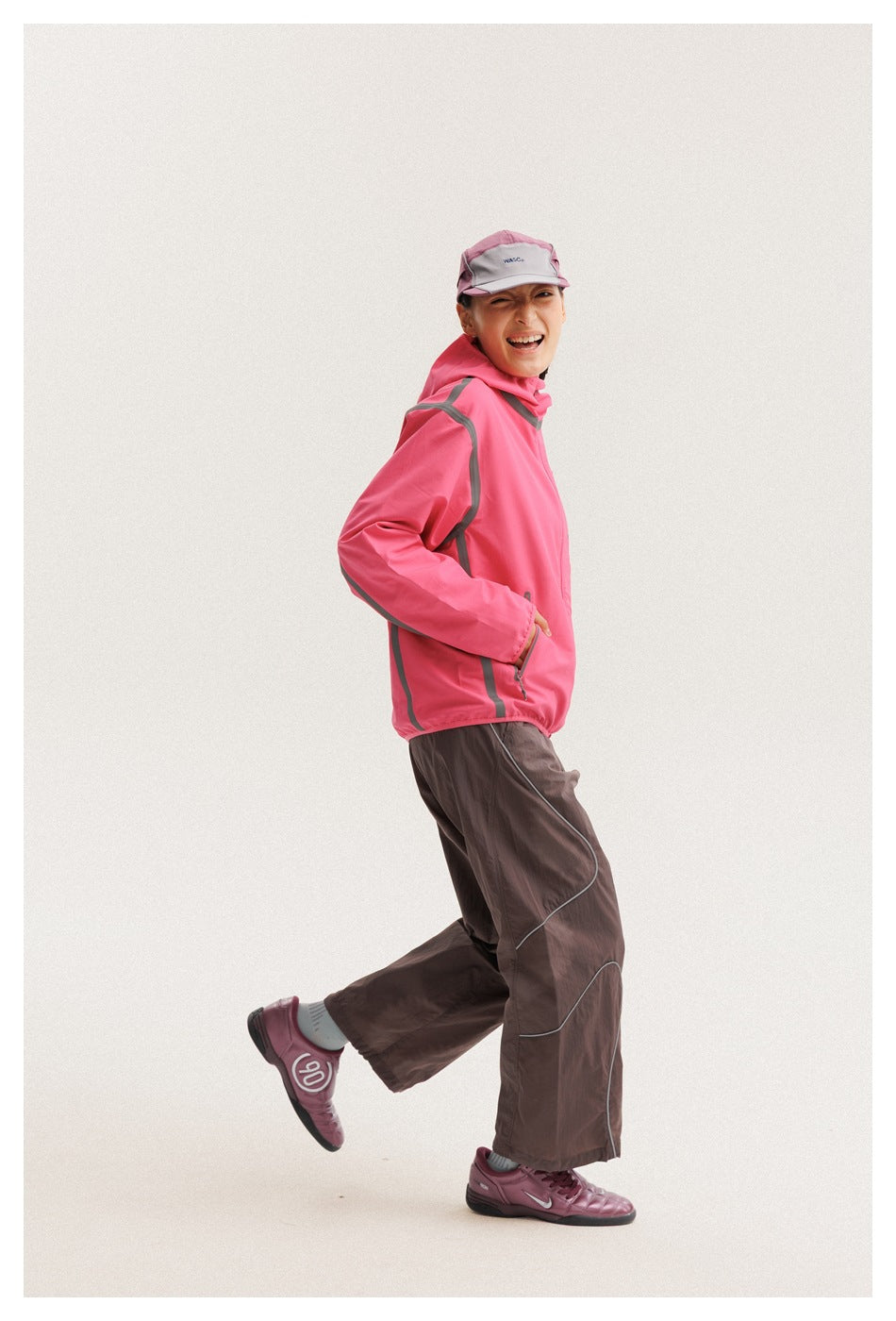 Model wearing a bright pink hooded jacket with gray trim and dark brown trousers, shown from the side.
