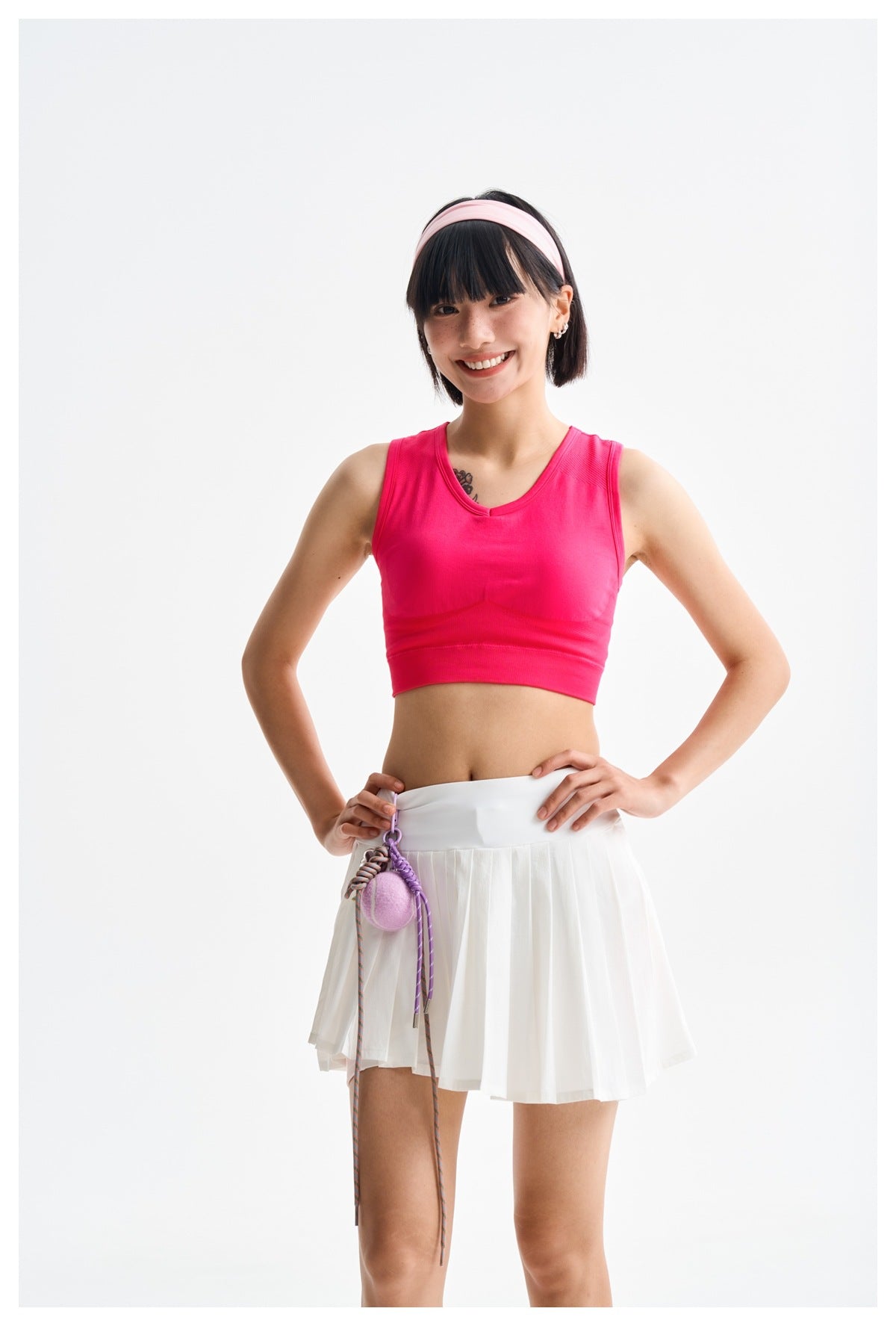 Smiling model wearing a hot-pink sleeveless crop top and white pleated tennis skirt with a wide waistband.
