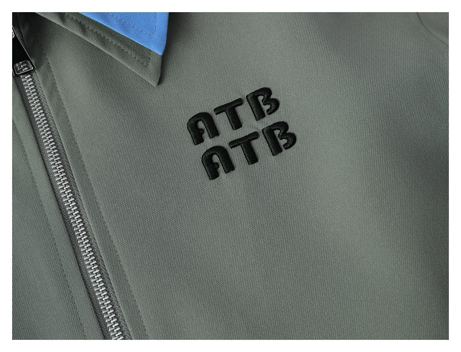 Close-up of a gray-green jacket with black embroidered lettering, a blue collar accent, and a metal zipper.
